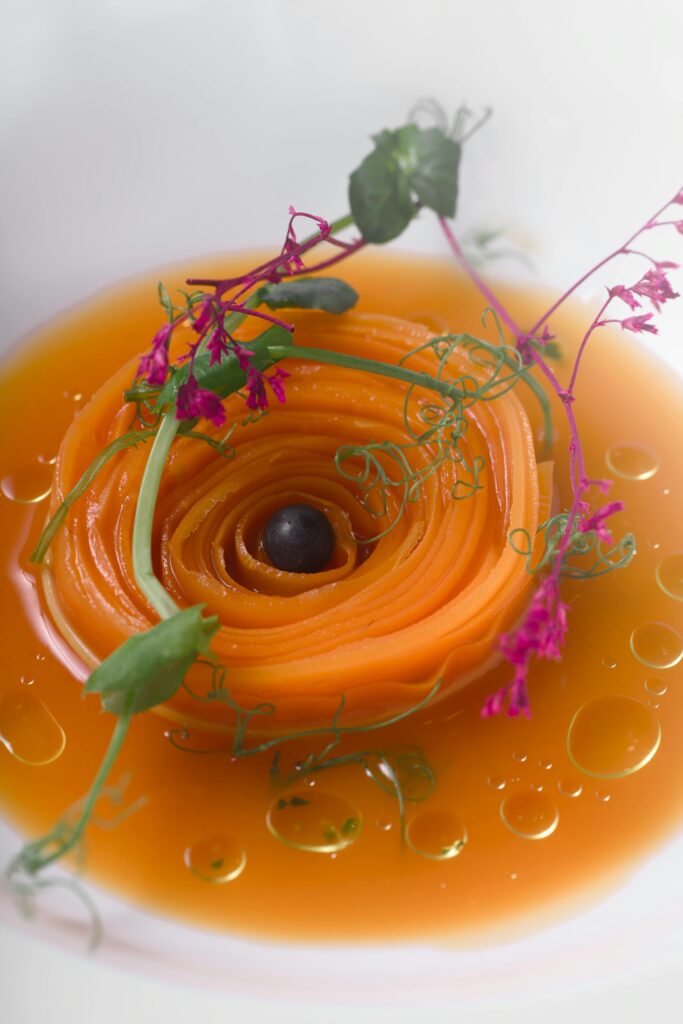 L'immagine mostra un antipasto a base di carote in forma di spirale, servita con guazzetto e fiori eduli. Piatto perfetto per i segni d'aria a tavola